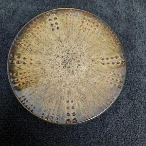 May include: A round ceramic plate with a brown and beige speckled glaze. The glaze has a textured, almost feathery pattern.