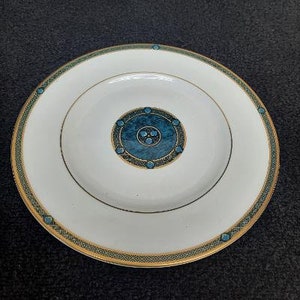 May include: White porcelain plate with a gold rim and a teal and gold geometric design. The plate features a central teal medallion with a circular pattern and teal accents. A decorative piece for dining.