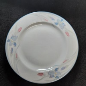 May include: A white ceramic saucer with a blue and pink floral design. The saucer has a scalloped edge and is perfect for serving tea or coffee.