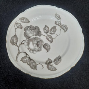 May include: A white ceramic plate with a gold rim features a gray and white floral design of roses and leaves.