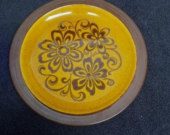 May include: A brown and yellow ceramic plate with a floral design. The flowers are brown and have a stylized design. The plate has a brown rim.