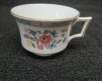 Può includere: Una tazza da tè bianca con un bordo dorato e un motivo floreale. La tazza ha un manico ed è decorata con fiori rosa, blu e verdi.