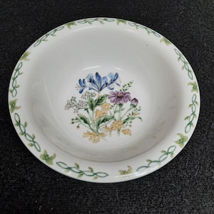May include: White ceramic bowl with a floral design. The bowl features a central bouquet of blue, purple, and white flowers with yellow and green foliage. A green vine and flower pattern decorates the rim.