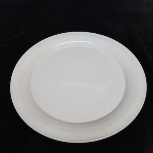 May include: A white ceramic plate with a raised center and a wide rim. The plate is round and appears to be unused. The background is a solid black color.