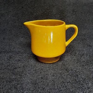 May include: A yellow ceramic creamer with a rounded body and a handle. The creamer has a textured base.
