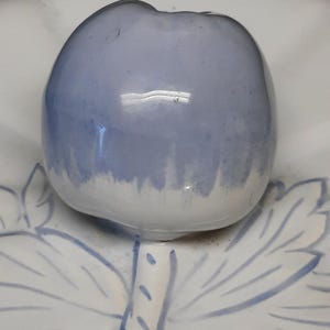 Puede incluir: Una tapa de cer&aacute;mica azul claro con forma de manzana, con una base blanca, se asienta sobre un plato blanco con dise&ntilde;os azules de hojas y tallos. La tapa de la manzana tiene un acabado brillante y un sutil degradado de color.