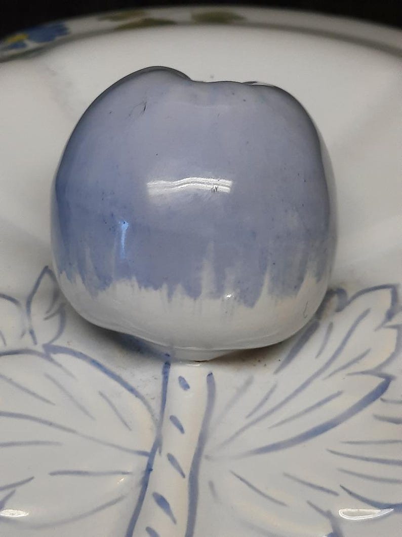 Puede incluir: Una tapa de cer&aacute;mica azul claro con forma de manzana, con una base blanca, se asienta sobre un plato blanco con dise&ntilde;os azules de hojas y tallos. La tapa de la manzana tiene un acabado brillante y un sutil degradado de color.