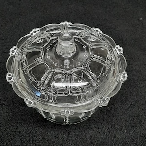 May include: A clear glass candy dish with a lid. The dish has a scalloped edge and a decorative pattern on the lid and base. The lid has a small knob for easy lifting. The dish is designed to hold small candies or other treats.
