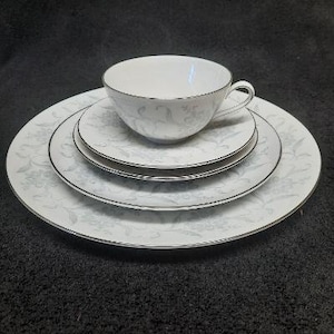 May include: A set of four white ceramic plates with a delicate floral pattern in shades of gray. The set includes a saucer, a teacup, and two larger plates.