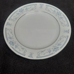 May include: A white porcelain plate with a delicate floral pattern in light blue and gray. The plate has a thin black line near the edge and a decorative border. The plate is round and has a smooth surface.