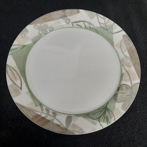 May include: A white ceramic dinner plate with a green, brown, and gray floral design. The plate has a slightly raised rim.