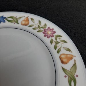May include: White ceramic plate with a black rim and a floral design featuring pears, flowers, and leaves. The design is in shades of pink, yellow, green, and blue.