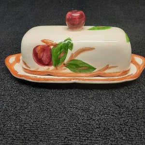 May include: A white ceramic butter dish with a red apple design. The dish has a lid with a raised apple on top. The dish is decorated with brown branches and green leaves.