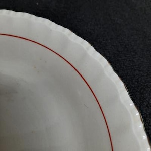 May include: White ceramic plate with a scalloped edge and a thin red line around the rim.