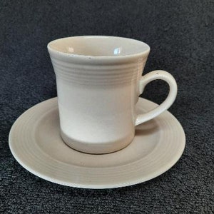 May include: A white ceramic coffee cup and saucer set. The cup has a simple design with a slightly flared rim and a handle. The saucer has a matching design with a slightly raised edge.