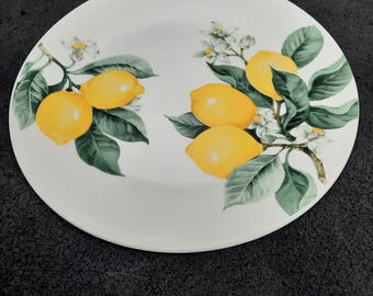 May include: White ceramic plate with a painted design of lemons, green leaves, and white blossoms. The lemons are yellow and the leaves are green. The plate is round and has a slightly raised edge. A decorative piece for the kitchen.