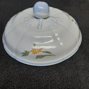 May include: White ceramic lid with a light blue knob. The lid features a floral design with orange and green flowers, and blue leaf accents. A blue stripe borders the edge.