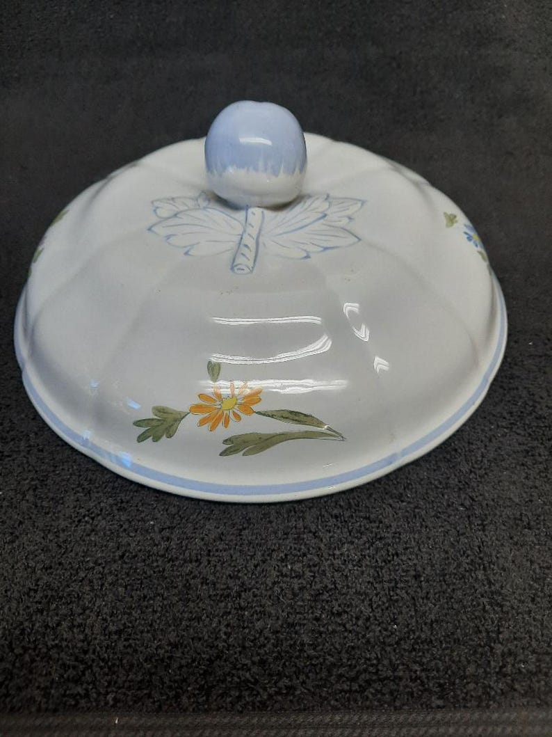 Puede incluir: Tapa de cer&aacute;mica blanca con un pomo azul claro. La tapa presenta un dise&ntilde;o floral con flores naranjas y verdes y detalles de hojas azules. Una franja azul bordea el borde.