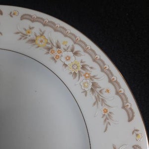 May include: A white ceramic dinner plate with a gold rim and a floral design in shades of brown, yellow, and white.