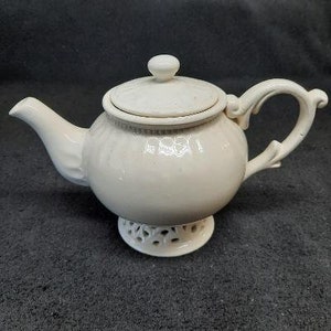 May include: White ceramic teapot with a lid and a decorative base. The teapot has a round body and a curved spout.