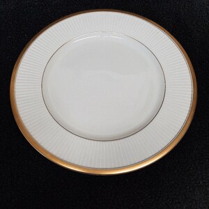 May include: A white ceramic plate with a gold rim and a textured, ridged pattern on the outer edge. The plate has a simple, elegant design, suitable for serving food. The plate is round.