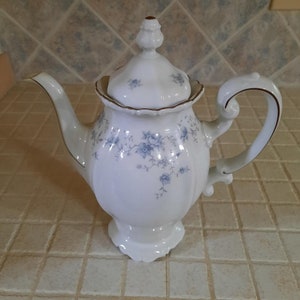 May include: A white porcelain teapot with a blue floral pattern and gold trim. The teapot has a lid with a knob on top and a curved handle.