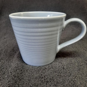 May include: A light blue ceramic mug with a ribbed design and a handle. The mug is sitting on a dark gray surface.