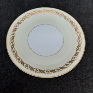 May include: A white ceramic saucer with a gold rim and a gold floral design around the edge.