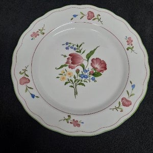 May include: A white ceramic dinner plate with a scalloped edge and a floral design. The design features pink, red, and yellow flowers with green leaves and blue accents.