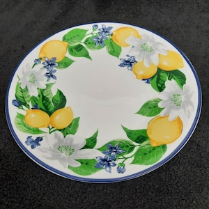 May include: White ceramic plate with a blue rim, decorated with a lemon and floral design. The design features yellow lemons, white flowers, green leaves, and small blue flowers arranged around the plate's edge.