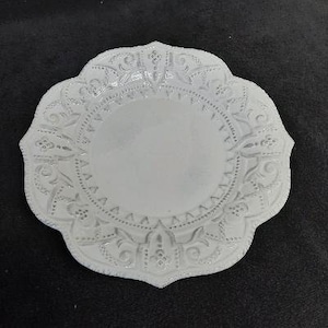 May include: White ceramic plate with a decorative, scalloped edge and an intricate floral design.
