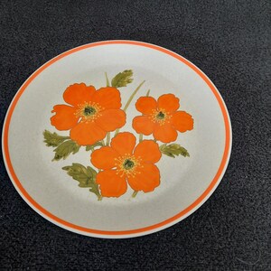 May include: Round ceramic plate with a floral design. The plate features three orange flowers with green leaves on a speckled, off-white background. An orange border encircles the edge of the plate.
