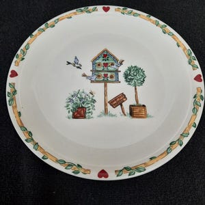 May include: A white ceramic plate with a green vine border and red hearts. The center of the plate features a colorful birdhouse with two birds, a small tree, and a sign that reads "Meet Me By The Garden".