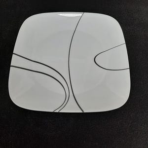 May include: White square ceramic plate with a black abstract design. The design features curved lines and shapes.