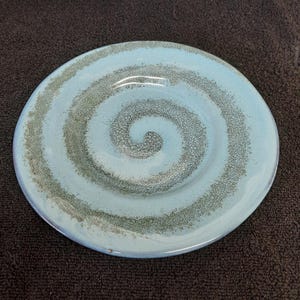 May include: A light blue ceramic plate with a swirling green design. The plate has a textured surface and a slightly raised rim.