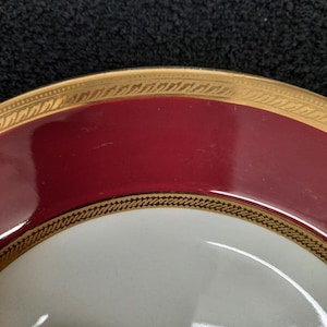 May include: Close-up of a red and white ceramic plate with a gold trim. The plate has a simple design with a gold band around the edge and a smaller gold band in the center.