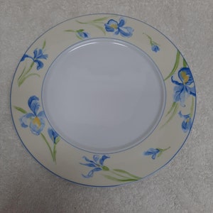 May include: A white ceramic dinner plate with a blue and green floral design. The plate features a border of blue irises with yellow centers on a cream background.