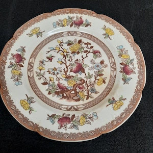 May include: A white ceramic plate with a brown, yellow, and red floral design. The plate has a scalloped edge and a brown border.