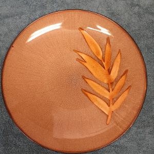 May include: A round, orange-brown ceramic dinner plate with a stylized leaf design in a lighter shade of orange.