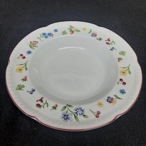 May include: White ceramic soup bowl with a red rim and a floral pattern around the edge. The pattern features red, yellow, blue, and green flowers, strawberries, and cherries.
