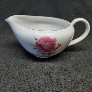May include: A white ceramic creamer with a single pink rose design. The creamer has a handle and a rounded shape.