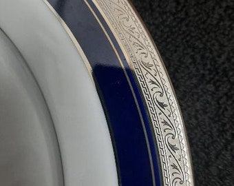 Op de afbeelding: Een wit keramiek bord met een blauwe en zilveren rand. De rand heeft een decoratief patroon van zilveren swirls en lijnen.
