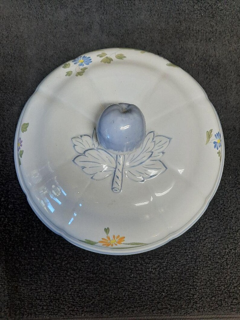 Puede incluir: Una fuente de cer&aacute;mica blanca con tapa. La tapa presenta un asa en forma de manzana azul claro y un dise&ntilde;o de hojas. El plato est&aacute; decorado con peque&ntilde;os detalles florales azules y amarillos.