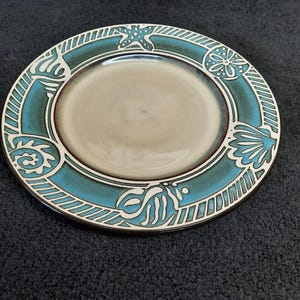 May include: A ceramic plate with a teal and white sea-themed border. The border features starfish, seashells, and other aquatic designs. The center of the plate is a light beige color.