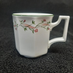 May include: White ceramic mug with a green rim and a pink and green floral design. The mug has a octagonal shape and a handle.