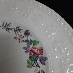 May include: White ceramic plate with a raised rim and floral design. The design features pink, green, and purple flowers with green leaves. The plate has a white background.