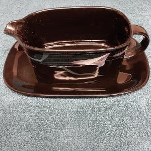 May include: A brown ceramic gravy boat with a matching saucer. The boat has a simple design with a subtle swirl pattern.