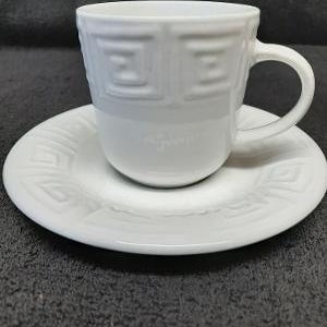 May include: White ceramic teacup and saucer set with a geometric pattern. The cup has a handle and the saucer has a raised edge.