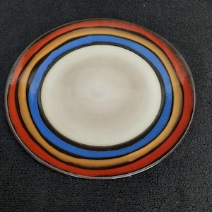 May include: A round ceramic plate with a white center and concentric rings of red, orange, brown, and blue.