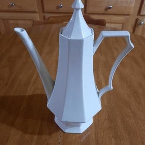 May include: White ceramic teapot with a lid and a handle. The teapot has a geometric design and a pointed top.